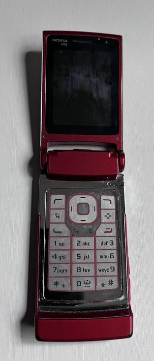 A red Nokia N76 flip phone, open to display its keypad and screen. The keypad features alphanumeric buttons, with some wear visible on the surface.