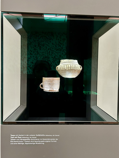 Vitrine, darin zwei Keramikgfäße. Nach links (vom Menschen vor der Vitrine aus) unten eine Henkeltasse mit etwa handtellergroßer Öffnung, die rundherum mit einem eingedrückten Punktmuster verziert ist.