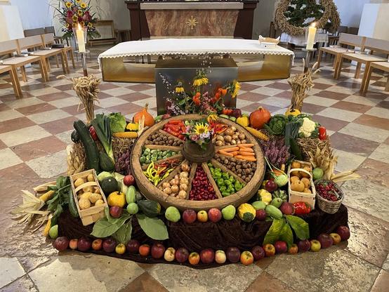 Blick in den Altarraum einer Kirche: Vor einem modernen, schlichten Volksaltar aus Edelstahl ist ein Erntedank-Arrangement aufgebaut. In der Mitte ist ein Wagenrad, zwischen den Speichen sind säuberlich und bunt Karotten, Nüsse, Rosenkohl, Zwiebeln, Tomaten etc. sortiert. Drumherum sind größere Früchte, Feldfrüchte und Gemüse aufgebaut und am Rand mit Herbstblumen geschmückt. Der Boden der Kirche ist schachbrettartig rot-weiß gemustert, ausgetreten und alt. Der Altar ist mit einem weißen Tuch bedeckt, rechts und links brennen auf Leuchtern zwei Kerzen. Im Hintergrund ist der Ansatz eines Hochaltars zu erkennen, einige Stühle und ein großer Blumenstrauß mit Sonnenblumen sowie eine große Erntekrone aus Getreide und Grünzeug.