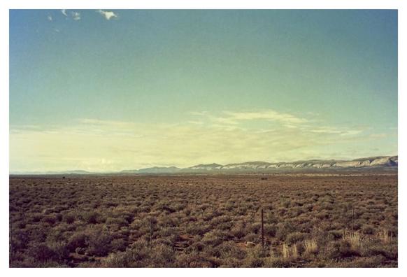 Expansive desert landscape under a vast blue sky. Distant mountain range on the horizon, with dry, sparse vegetation covering the foreground. Calm and serene.