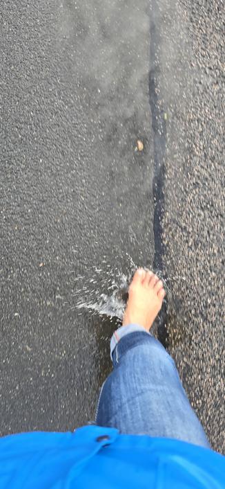 Das bild zeigt von oben , wie ein Mann bei Regen durch eine Pfütze läuft. Das Hosenbein der Jeans ist hochgekremüelt und das Wasser spritz zur Seite weg, während er in die Pfütze tritt.