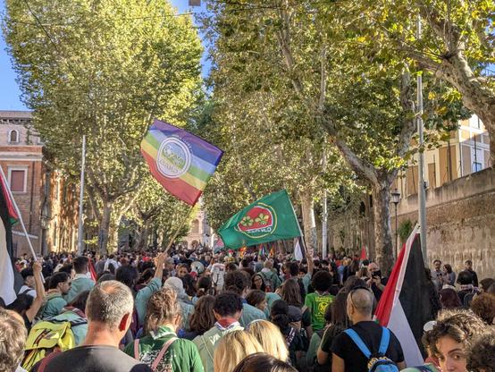 Il corteo di manifestanti avanza lungo un viale alberato. Sopra le loro teste si vedono due bandiere dell'associazione scout CNGEI.