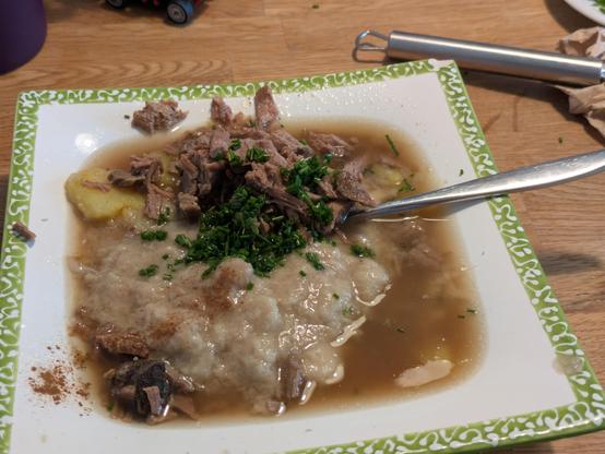 Das Bild zeigt ein Gericht mit Suppe, das auf einem quadratischen, hellgrünen Teller mit einem verzierteren Rand serviert wird. Die Suppe hat eine hellbraune Farbe und enthält verschiedene Feststoffe, darunter große Stücke Fleisch, kleine weiße Bohnen und eine zerkleinerte, dunkle grüne Kräuterstreuung, die in der Mitte platziert ist. Ein Silberschalenlöffel ragt in die Suppe hinein und scheint die Textur der Suppe zu verteilen.