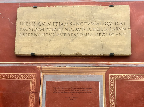 Auf falschen Marmor angebrachte lateinische Inschrift. Darunter die deutsche Übersetzung: "Sie glauben sogar, dass den Frauen etwas Heiliges und Sehendes innewohnt, und deshalb weisen sie weder ihre Ratschläge ab, noch lassen sie ihre Weissagungen unbeachtet." (Tacitus, Germania 8.2, circa 98 nach Christus)