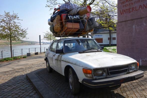 A weathered, white vintage Saab car, parked on a cobblestone surface near a river. The car is heavily loaded with an assortment of luggage, bags, and household items strapped precariously to its roof, including a bag with the word ‘YEAH’ written on it. The items also include a large wicker basket, a rolled-up rug, a wooden chair, and various bags, all secured with ropes. The car appears dusty and well-travelled, with visible rust and wear. In the background, a pink monument or signpost with the word ‘VATERLAND’ is visible, alongside a serene riverside view with trees and buildings in the distance.