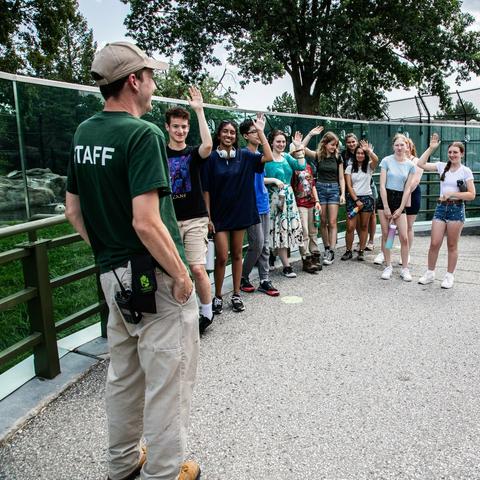 In the image, a man dressed in a green shirt and baseball cap is standing on a paved area, waving to a group of people who are gathered around him. There are at least eight individuals in the scene, some of whom are waving back, while others stand in a line or group formation.

In the background, there is a fence visible, adding to the outdoor setting of the image. The man's position relative to the fence suggests that he might be a zookeeper or a staff member from a park or recreational facility.