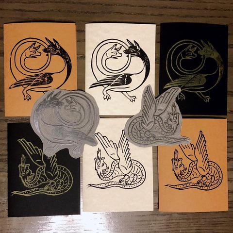 Six block print cards laid out under their carved blocks, two each of parchment pattern, kraft brown and black. there are two prints: one of a dragon standing it’s own tail like a platform spouting fire straight up with unfurled feathery wings and the other of a creature with two heads, wings and two legs with paws, one head is situated at the end of a long tail that is forming a spiralling circle up and over the main body, the ’main’ Head is biting the tail as it circles near it and the second head is looking at it in shock/outrage/dispair