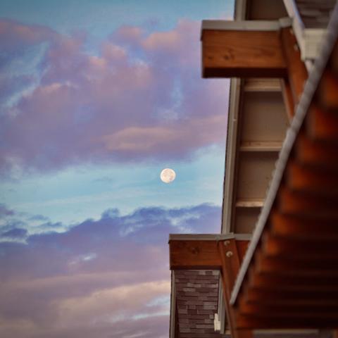 A nearly full moon peeks through horizontal clouds that frame it, as the wooden roof of a building is visible to its right in the foreground on a cool-colored morning.