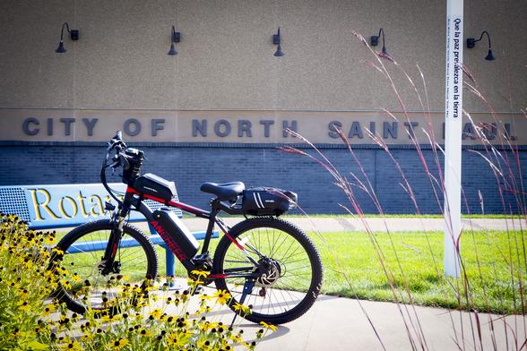 A black and red electric bike parked in front of a blue bench that says "Rotary" in gold letters. A wall in the background says "CITY OF NORTH SAINT PAUL"