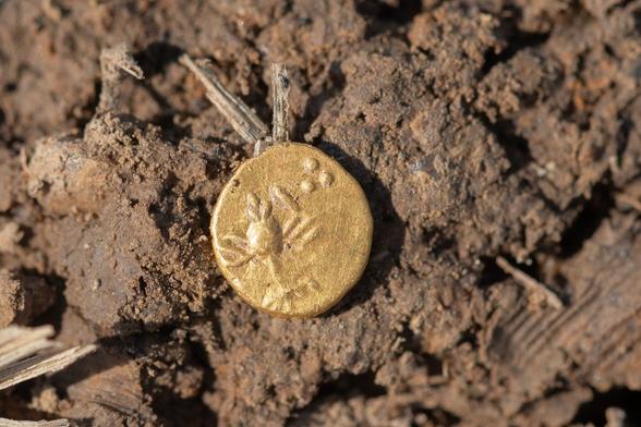 Moneta d'oro antica parzialmente affiorante dal terreno, con un disegno visibile sulla superficie, circondata da terra e rametti.