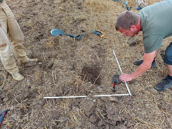 Due archeologi impegnati in scavi in un terreno agricolo, con attrezzature da scavo e un quadrato tracciato nel suolo per la mappatura.
