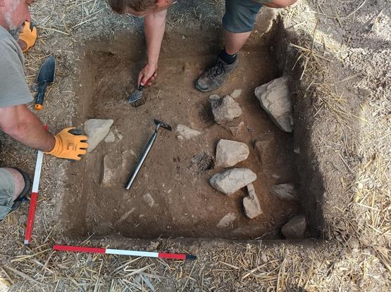 Due archeologi mentre scavano in un sito archeologico, circondati da rocce e strumenti da scavo, in un'area di terreno terrazzato.
