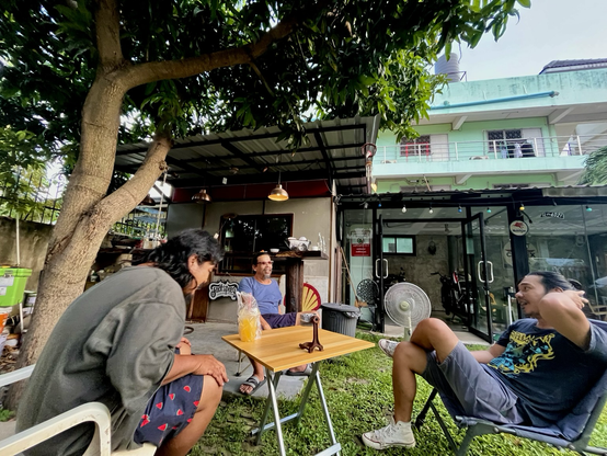 Three Thai men sit together under a mango tree in front of a glass room with a triumph motorcycle inside. The man in the right wears a parliament funkadelic shirt, the man in the left wears watermelon shorts. A Shell gasoline and a Fuck Speed sign can be seen.
