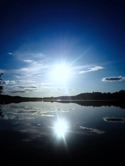 A serene lake reflecting a bright sun and blue sky, with scattered clouds and a peaceful shoreline in the background.