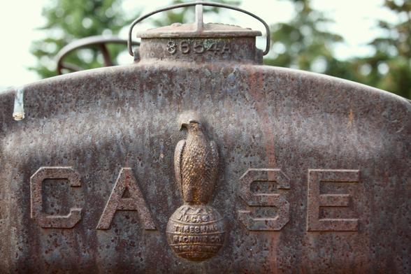 Close-up photo of the front steel grill of an old Case tractor/thresher machine. It is rusted and weathered and has bird shit on it, which is ironic since the eagle known as "Old Abe" is the centrepiece of the raised Case logo, sitting atop a globe between the CA and the SE, staring stoically off into the distance.

The words on the globe are: "J.I. Case Threshing Machine Co. Racine, Wis, U.S.A"

Visible above the grill is what looks like a large valve opening with a handle and the numbers, which may be "3654A" although it is hard to say for sure.