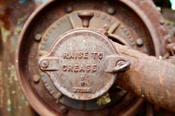 A close-up of part of the thresher/tractor which looks like a valve with a key to open or loosen it. It is rusted and the raised words on it are "RAISE TO GREASE" with a mini version of the eagle mascot Old Abe and a model number of 3718A.