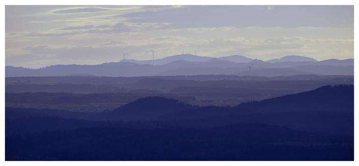 Long-distance shot of the Black Forest and the hills in the foothills, which glow in bluish light in varying contrasts. Several wind turbines and a transmission tower are visible on the Black Forest, standing out against the sky on the horizon.