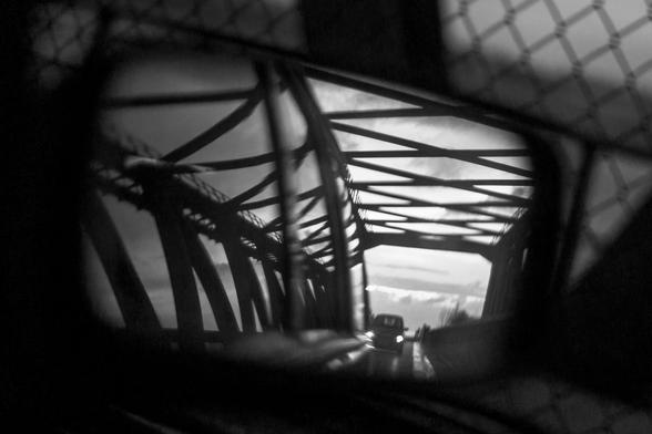 Schwarzweiß. Blick in einen rechten Rückspiegel eines Autos. Darin ist die Konstruktion einer Stahlbrücke zu sehen. Auf der Straße fährt ein Auto mit beleuchteten Scheinwerfern. Alles wirkt düster und dramatisch, auch wenn der Himmel im Hintergrund noch hell ist. Es könnte eine Verfolgungsszene aus einem 60er-Jahre-Krimi sein.
