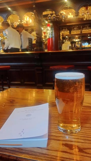 Looking past a pint of golden ale and a white, paperback book to the dark, carved wooden island bar of the Abbotsford pub