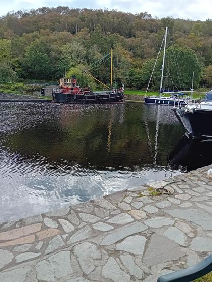 View of Crinan Basin with an old Clyde Puffer boat moored at the far side. The boat has a black hull and red and dark yellow cabin with a red and black funnel and yellow mast. To the right is a smaller sailing boat and the bow of a modern cruiser. Behind the basin is a bank of woodland and in the foreground a stone paved path.