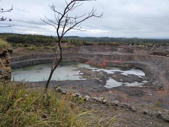 Inna odkrywka, widoczna ze skarpy z jedyną przeszkodą w postaci łysego drzewka pośrodku. Tu woda jedynie częściowo zalewa otwór, w jego dalszej części. Bliższa jest pokryta czarną ziemią, miejscami czerwoną, w wielu miejscach gęsto pokrytą szarymi kamieniami. Owa ziemia zachodzi na jeziorko, rozprzestrzeniając się półwyspem w kształcie nierównej litery "T". Centralny otwór otaczają trzy kolejne, szerokie półki, ze stromymi, czarno-szarymi ścianami. Miejscami ze ścian osuwają się stosy szaro-brązowych kamieni; po przeciwległej stronie jeziorka takie osuwisko otacza sporo czerwonej ziemi. Po prawej stronie widać maszyny, budowle, zwałowarki. W tle kolejne warstwy zielonego lasu, tworzące pofałdowany krajobraz. Niebo pochmurne, srebrnobłękitne.