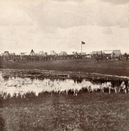 View of Camp Fisk, a temporary camp at St. Cloud, Minnesota for emigrants gathering to leave for Montana Territory as part of the Fisk Expedition of 1866. The camp was named for expedition leader, Captain James L. Fisk. Visible are many canvas tents, men, horses and/or mules, and many covered wagons. An unidentified flag flies over the camp and a pond or small lake is in the foreground. Possibly #4 in the series: 'Crossing the Plains to Montana, with Capt. Fisk’s Expedition, 1866.'