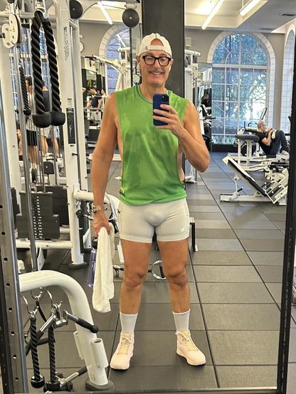 A fit individual (me) is taking a selfie in a gym mirror. They are wearing a green sleeveless shirt and light grey shorts. The person holds a towel and a water bottle, and they have on pink athletic shoes. Gym equipment is visible in the background.