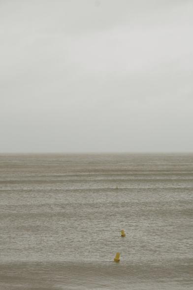 A tranquil seascape unfolds under a heavy gray sky, the muted tones of water and air blending seamlessly into one another. The photograph’s composition is striking in its minimalism and restraint, with the frame divided almost evenly between the vast, pale sky and the calm, textured sea. This balance draws attention to the horizon line—a thin, almost imperceptible boundary that anchors the image. Gentle, rhythmic waves introduce a subtle sense of movement, while a diagonal line of small yellow buoys punctuates the scene, guiding the viewer’s gaze from the foreground toward infinity. Their bright color contrasts softly against the subdued palette, serving as both visual anchors and narrative hints of human presence. The interplay of tone, texture, and space creates a meditative atmosphere, where stillness, simplicity, and balance become the photograph’s quiet strength.