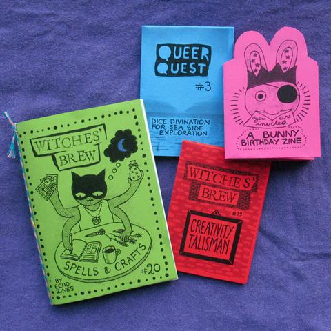 4 zines on a lavender-indigo coloured fabric. The zines have green, blue, pink, and red cover pages and are printed with black ink. The zine titles are Witches' Brew 19 and 20, Queer Quest 3, and You're Invited. The latter has a drawing of a bunny with a crown and an envelope in its mouth on the cover. The green zine features a witchy cat.