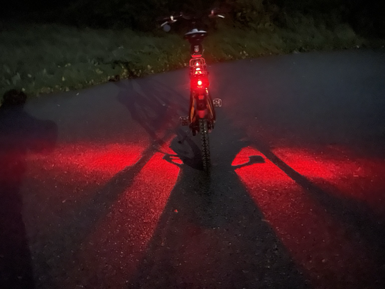 Foto eines Fahrrades von hinten. Ein Rücklicht scheint ganz normal, ein zweites ist so eingestellt, dass es links und rechts breite rote Lichtstreifen auf die Fahrbahn projiziert.