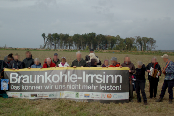 Eine Gruppe Menschen steht auf einer Wiese und hält ein Banner mit zahlreichen NGO-Logos und Aufschrift: "Braunkohle-Irrsinn. Das können wir uns nicht mehr leisten"; im Hintergrund die Laubbäume des besetzten Sündenwäldchens