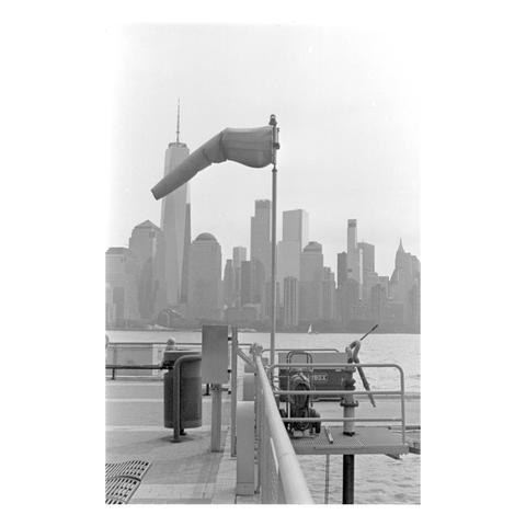 A windsock on a pole in the foreground, pointing left, against the backdrop of the Lower Manhattan skyline across the water. The windsock is partially inflated, indicating a gentle breeze.