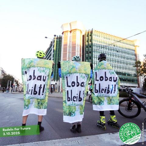 Drei Personen auf der Starße vor dem Verkehrsministerium. Alle drei haben ein Banner #LobauBleibt umgehängt.
