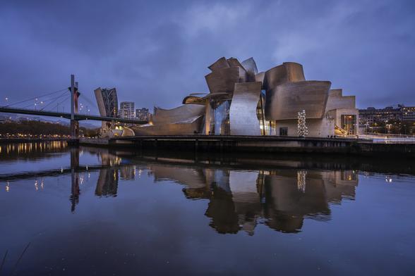 Reflected view of Bilbao's Guggenheim Museum on the Nervion river on a cloudy evening at blue hour. Designed by Frank Gehry, the museum’s structure is a complex interplay of abstractly curved forms, evoking a sense of ordered chaos. Although the metallic form of the exterior looks almost floral from above‭, ‬from the ground the building more closely resembles a boat‭, ‬evoking the past industrial life of the port of Bilbao‭. ‬Constructed of titanium‭, ‬limestone‭, ‬and glass‭, ‬the seemingly random curves of the exterior are designed to catch the light and react to the sun and the weather‭.