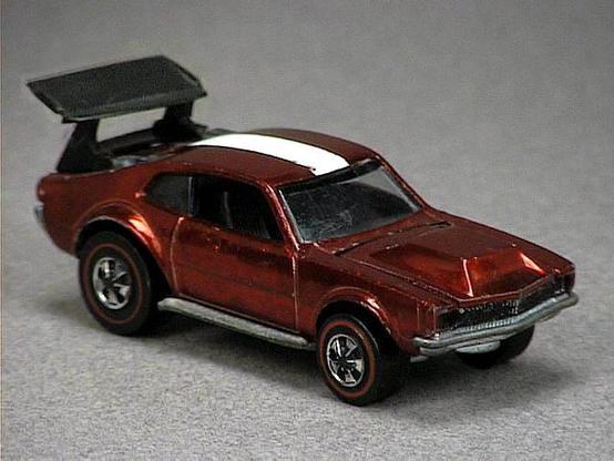 Released in 1970, Spectraflame Red Mighty Maverick of 1970 Hot Wheels with Redline wheels. Notes: Hood lifts up to reveal a metal engine. comes with a black or white stripe on roof, black plastic rear spoiler. HK version has a thinner stripe on the roof and unpainted headlights. Packaging: 1970 Blister. Metal Body, Metal Base. Made in Hong Kong. Scarcity: 5/10