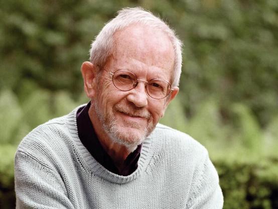 A close-up portrait of a person with gray hair and glasses, smiling gently at the camera. The person is wearing a dark-colored collared shirt under a light gray, knitted sweater. They have a warm complexion and visible wrinkles around their eyes and mouth. The background is a blurry green, appearing to be foliage or shrubbery. The person is looking directly at the viewer with a soft expression.

Provided by @altbot, generated privately and locally using Gemma3:27b