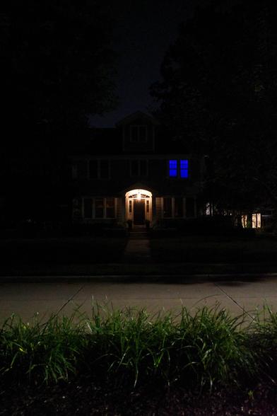 Color photograph looking across a well-lit residential street at night at a large house that's set back from the brightly-lit street, bright porch light in an arched doorway standing out from the darkened house that recedes into the darkness of trees in the background. Deep blue light shines through an upstairs window