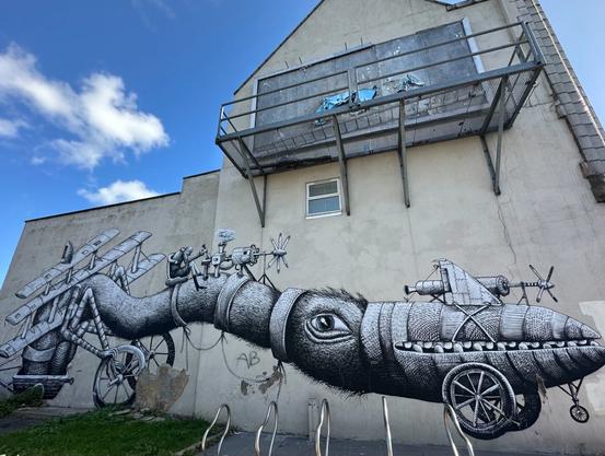 a large, highly detailed black and white mural on a wall depicting a fantastic creature/contraption that’s like a biplane built around an ostrich-like bird. A humanoid figure is seated on a chair strapped to the neck.