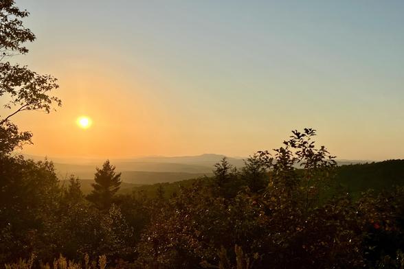 A scenic view of a sunset over southern New Hampshire mountains.  Viewed from a rocky opening in the forest. There are trees in the foreground, lit by a warm, golden light across the descending landscape. The sky transitions from orange to blue as the sun descends on the horizon.