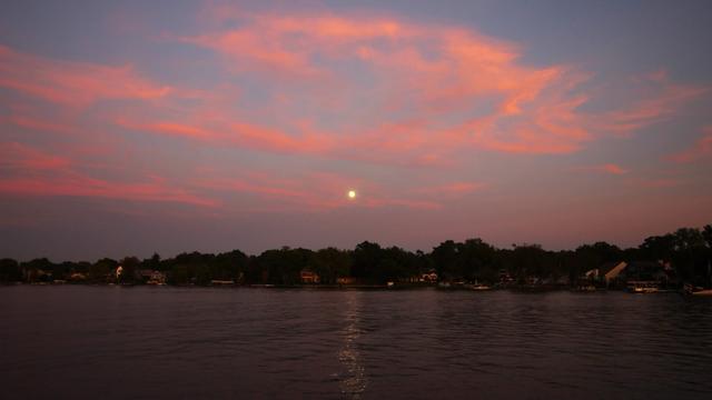 Photo of a nearly full moon rising over a lake surrounded by pink clouds.