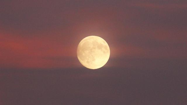 Photo of a medium-large nearly full moon surrounded by pink clouds.