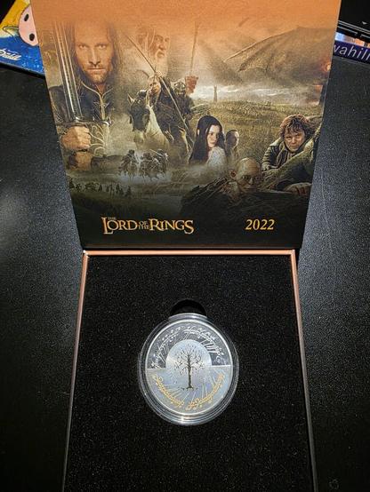 A collector’s display box featuring a 2022 commemorative silver coin from Malta, themed around The Lord of the Rings. The presentation box is open, showing the richly illustrated lid with warm golden and brown tones showing iconic characters and scenes from the movie trilogy — including Aragorn holding his sword, Gandalf on horseback, Arwen with her long dark hair, Frodo and Gollum in the foreground, and a vast battle scene in the distance beneath a cloudy sky. The Lord of the Rings logo and the year “2022” appear in gold lettering near the bottom of the backdrop. In the black velvet-lined base, the coin is securely encased in a clear protective capsule. The coin itself gleams in silver, depicting the White Tree of Gondor at its center with elegant Elvish Tengwar script engraved along the upper and lower edges. The combination of silver detailing and subtle golden inscriptions gives the piece a refined, almost mythical appearance, celebrating the fantasy legacy of Tolkien’s world.