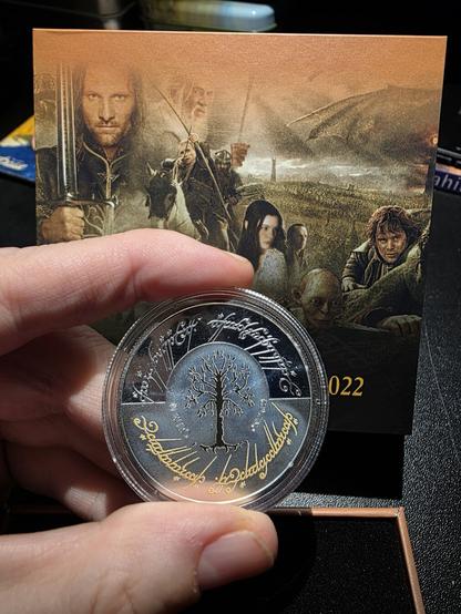 A close-up photo showing the Lord of the Rings 2022 silver coin from Malta, carefully held between two fingers after being lifted from its display box. The coin, still encased in a clear protective plastic capsule, gleams under the light, revealing the detailed design of the White Tree of Gondor at its center — a symbol of hope and renewal in Middle-earth. Surrounding the tree is a crescent-like silver glow, giving the impression of moonlight shining on the emblem. Along the upper and lower edges, fine Elvish Tengwar script is engraved; the top inscriptions are in bright silver, while the bottom lines shimmer in gold, adding a regal contrast to the design. The coin’s surface alternates between matte and mirror-polished finishes, emphasizing its intricate craftsmanship. Behind it, the backdrop from the collector’s display remains visible, featuring iconic Lord of the Rings characters.