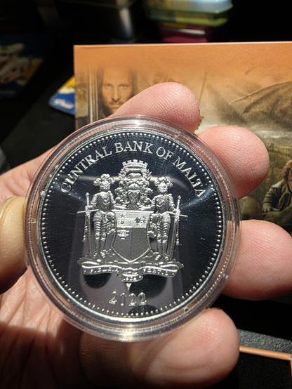 Close-up of a hand holding the reverse side of a 2022 silver coin from Malta, securely encased in a transparent protective capsule. The coin gleams under direct light, revealing the intricate design of the official Central Bank of Malta emblem engraved in high detail. At the top, the words “CENTRAL BANK OF MALTA” curve elegantly along the rim. Below, the national coat of arms is depicted: two armored knights standing guard on either side of a central shield, each holding a spear and wearing ornate helmets with plumes. The shield bears symbolic Maltese heraldry — including a small cross — and is crowned with a fortress-like crown above. The base of the design includes a ribbon scroll and the year 2022 finely engraved beneath it. The background is a mirror-like polished silver surface, contrasting beautifully with the frosted relief of the design. Behind the coin, partially blurred but still recognizable, is the orange-toned Lord of the Rings collector card with several characters.