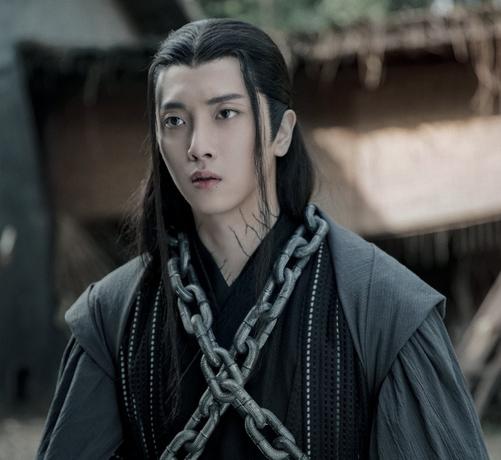 Close in shot of Wen Ning, clad in robes of grey and black with stout chains wrapped in an X around his torso. His expression seems concerned.