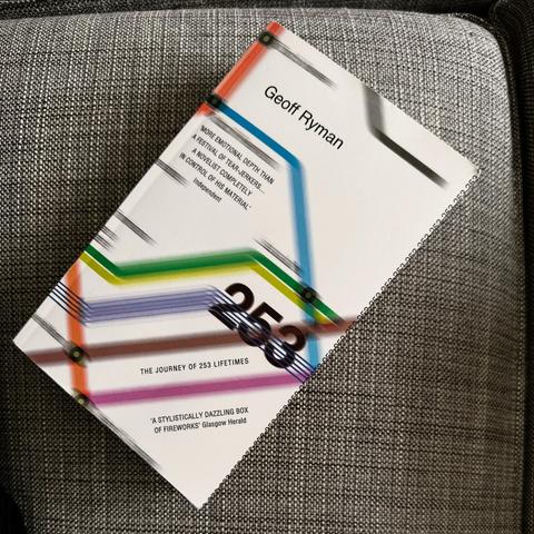 A book titled "253: The Journey of 253 Lifetimes" by Geoff Ryman is displayed on a textured gray surface. The cover features colorful graphic elements, including horizontal lines in various colors reminiscent of the map of the London Underground, and the prominent number "253."