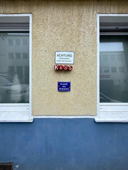das bild zeigt einen teil einer fassadenansicht eines gebäudes mit zwei fenstern. die fassade ist in einem sandfarbenen ton gehalten, der untere bereich ist blau gestrichen. zwischen den fenstern – das linke ist angeschnitten am linken bildrand und das rechte fenster ist angeschnitten am rechten bildrand – befinden sich drei schilder. das oberste weiße schild mit schwarzem text trägt die aufschrift "achtung liebespaare bitte hier küssen". darunter befindet sich vier rot bemalte konservendosen mit der gemeinsamen aufschrift "kiss". das unterste schild ist etwas kleiner und blau mit weißem text. es trägt die aufschrift "place des bisous" mit kleinen lippen-symbolen. die fenster reflektieren gebäude auf der gegenüberliegenden straßenseite und autos.