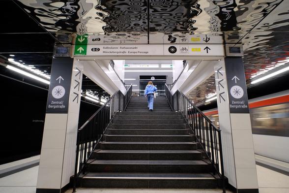 Fast symmetrisches Bild von einem Treppenaufgang, auf der eine blau gekleidete Person nach oben geht. Rechts Bewegungs-unscharf eine abfahrende U-Bahn mit rotem Streifen. Die Decke wellig verspiegelt wie eine Wasseroberfläche