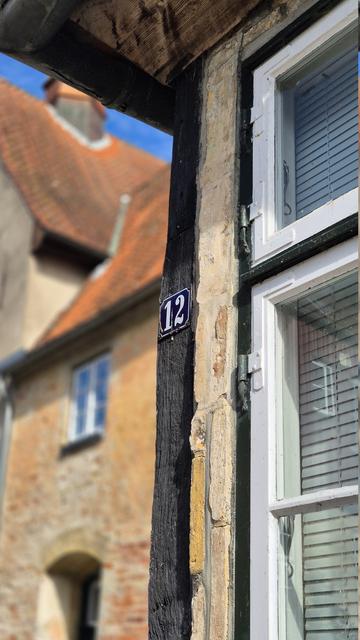 Im Zentrum des Bildes ist ein kleines, dunkelblaues Emailleschild mit weißem Rand und der Zahl 12 zu sehen. Es ist außen an der Fassade links neben einem Fenster an der Außenecke eines Hauses an einem Fachwerkbalken angebracht. Links, zurückversetzt, ist unscharf ein weiteres Gebäude zu erkennen.