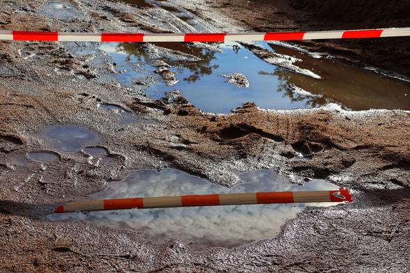 A close-up photograph of a waterlogged construction site. Large puddles of standing water reflect the sky, surroundings, and the red-and-white striped barrier tape, creating the illusion of a second tape. The uneven, muddy ground is cordoned off with the tape, indicating restricted access for safety reasons.